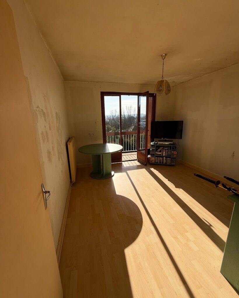 Salon lumineux avec parquet, table ronde verte et porte-fenêtre ouverte sur un balcon ensoleillé.